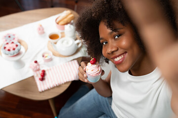 Beautiful young African blogger presenting piece of cupcake in concept special cuisine with selfie...