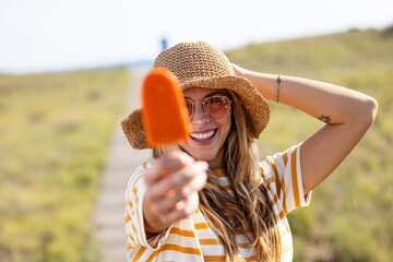 Attractive young woman eating a orange popsicle looking at the camera on summer - 790400391