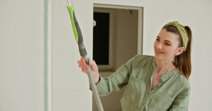 Young happy woman painting interior wall with paint roller in new house. Woman paints the wall. Portrait of a young woman making repairing in apartment. Painting inside new house