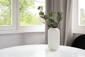 Background of white vase with artificial eucalyptus on white table and window at the back