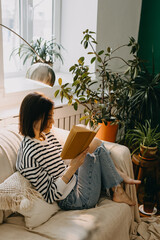 Young brunette woman reading a book on a couch, in soft day light by a window, surrounded by green...