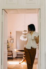 Woman savoring coffee at home, in a sunlit cozy room, standing in a doorway