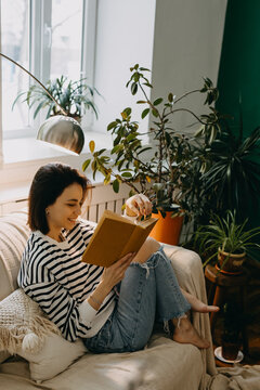 Young happy woman reading a book on a couch, in soft day light by a window, surrounded by green houseplants.