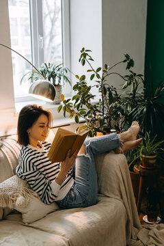 Young brunette woman reading a book on a couch, in soft day light by a window, surrounded by green houseplants.