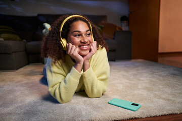 Cheerful young afro woman listening to music with headphones lying on carpet at home in evening. Latin girl using mobile phone enjoys favorite song in living room floor. Leisure time in generation z 