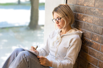 Blonde girl in glasses sits near the window and writes notes in a notebook. Beautiful woman pensively writing with pen in notebook, student, freelancer, cafeteria, sunny day, panoramic window