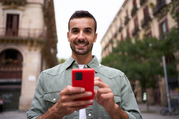 Portrait young European man stands looking smiling camera holding mobile phone on street in Madrid...