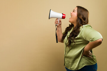 Profile view of a cute overweight woman using a megaphone in a studio with a beige backdrop - 790416323