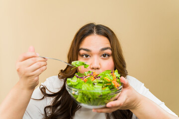 Cheerful young plus-size woman eating a healthy bowl of salad against a neutral background