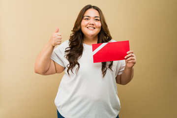 Pretty Hispanic plus-size woman in a photo studio holding a red gift card and giving a thumbs up sign - 790416571