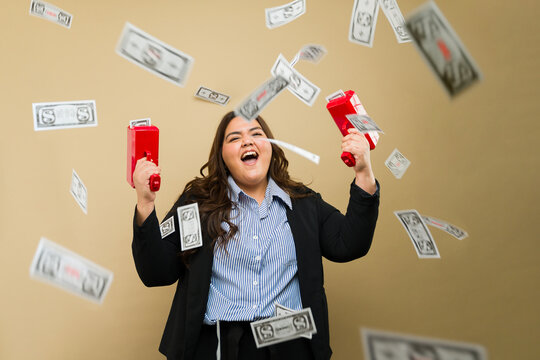Happy plus-size woman laughing and using a money gun to shoot bills on the air in a studio background