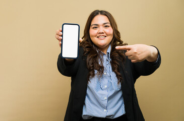 Smiling plus-size lady in business attire pointing to blank phone screen in studio - 790417317