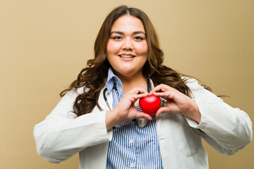 Cheerful plus-size female doctor smiling and holding a red heart in a studio - 790417523