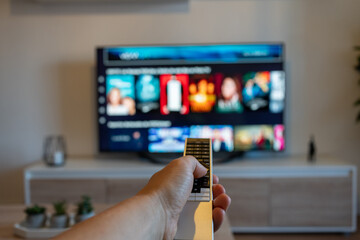 Hand of unrecognizable person holding remote control with television in front. Changing channels. Choosing and preparing a series on an online movie platform.