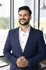 Happy confident Indian company CEO man in formal suit standing in office with hands folded, looking away, smiling, thinking on successful business ideas, management strategy, leadership