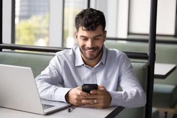 Happy successful young Arab freelance worker using digital gadgets in co-working space, enjoying online job communication, texting message on cellphone on corporate chat, smiling
