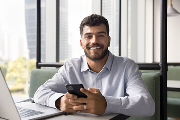 Happy young Arab freelancer man using gadgets for online work communication at co-working workplace, looking at camera with toothy smile. Entrepreneur, manager posing for business portrait