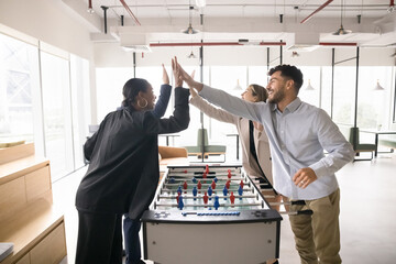 Happy diverse coworkers giving high five over table soccer field, celebrating win, enjoying recreation, leisure time on work break, group activity, playing toy football in modern co-working space