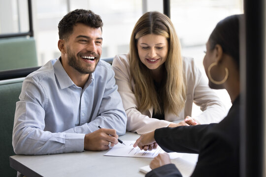 Happy married couple signing real estate property buying agreement, writing in document, meeting with broker, banker, agent at table, talking to consultant, smiling, laughing
