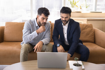 Two Indian and Arab business men watching online content on laptop, sitting on office couch, talking computer, discussing job application, service for work Internet communication