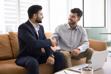 Positive Indian consultant and satisfied Arab client men shaking hands at laptop, sitting on office couch, meeting for consultation, getting agreement, enjoying professional communication