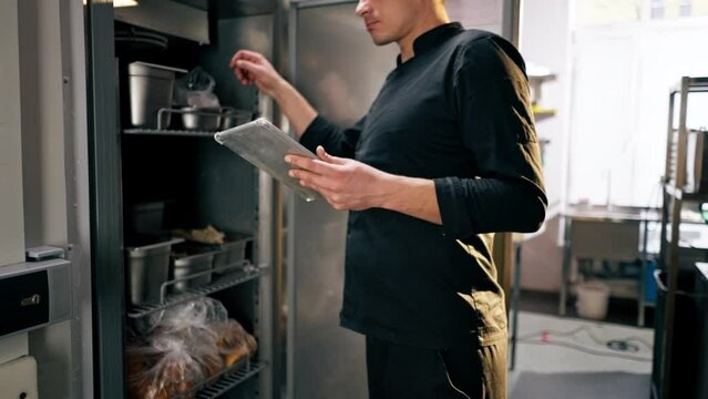 chef stands in a professional kitchen in a black uniform and looks through what is on the list in the refrigerator