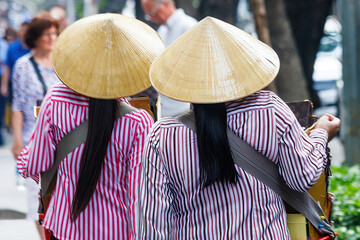 2 women walking down a street in Hanoi, Vietnam wearing conical hats - obrazy, fototapety, plakaty