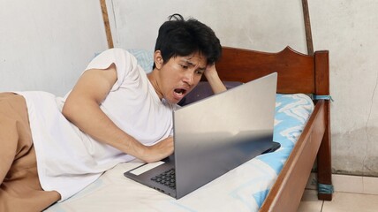 shocked young asian man lying on bed while watching movie on laptop