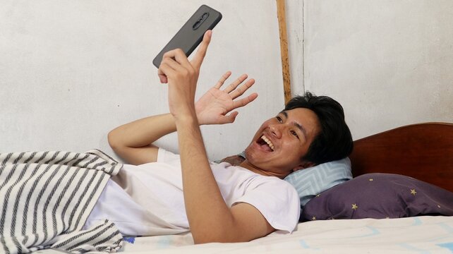 young Asian man lying on the bed in the bedroom playing on his smartphone