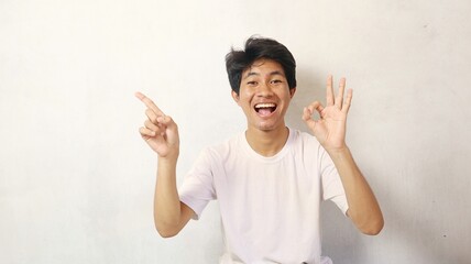Young Asian man poses happily while pointing to the side and gesturing okay on a white background