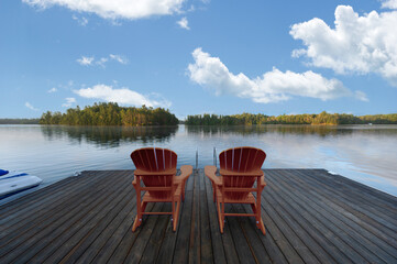 Two brown Adirondack chairs on the dock of a cottage greet the serene Muskoka summer morning,...