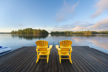 Two yellow Adirondack chairs on a wooden dock welcome the serene Muskoka morning, overlooking the...
