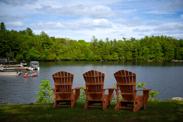 Naklejka premium Three Adirondack chairs on a lush green lawn overlook the tranquil blue waters of a lake. Two people paddle away on a canoe from the dock, infusing the serene scene with a hint of adventure.
