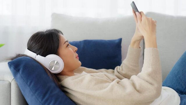 Happy young asian woman wearing headset and relax on comfortable couch at home texting messaging on smartphone, smiling girl use cell phone video call