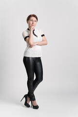 Business woman wearing white short sleeve blouse, black leather tight trousers and high heeled shoes looking at camera with one hand raised to chin, standing legs crossed at ankle on white background