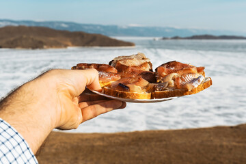 Sandwiches with omul on a white round plate. A plate of sandwiches in hands. Winter Baikal in ice.