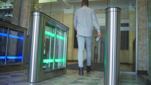 African American man or businessman walks through gates into business center using electronic pass card. Turnstile with touch panel for automatic scanning and identification in modern office building.