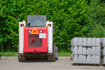 Pallets with a curb stone stacked on them and a red and white excavator stand on an asphalt road against a background of dense greenery