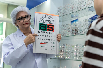 Old optician woman holding snellen number eye chart in optical clinic