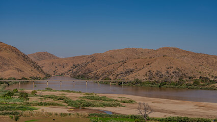 A road bridge passes over the red-brown river. Green vegetation on sandy shores. Red soil hills against a clear blue sky. The landscape of Madagascar. Tsiribihina river.