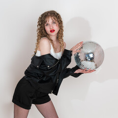 Confident woman is holding disco mirror ball, looking at camera. She's young adult pin-up girl from Generation Z era, with long curly hair. She dressed in clubbing style, standing on white background