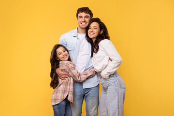Healthy family relationship. Loving parents and daughter embracing, standing over yellow studio...