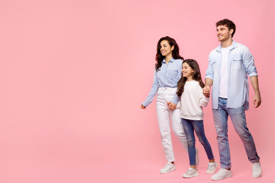 Young happy parents with daughter in casual clothes hold hands walking, going strolling isolated on pastel pink background, free copy space