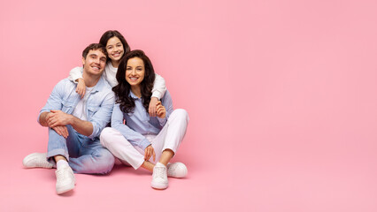 Parents and daughter sitting on pink background, posing together and smiling, girl embracing father and mother from back. Panorama shot with free space