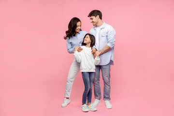 European parents embracing their happy daughter looking at pretty girl, standing together on pink studio background, full length shot