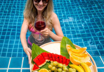 Beautiful woman in the swimming pool with glass of wine and floating tray of fruits - 790601975
