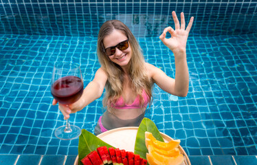 Beautiful woman in the swimming pool with glass of wine and floating tray of fruits - 790602356