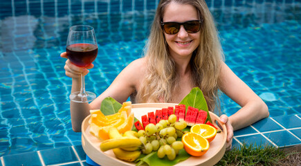 Beautiful woman in the swimming pool with glass of wine and floating tray of fruits - 790602545