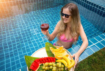 Beautiful woman in the swimming pool with glass of wine and floating tray of fruits - 790603782