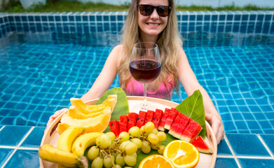 Beautiful woman in the swimming pool with glass of wine and floating tray of fruits